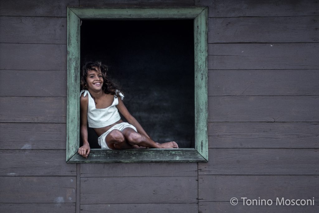 Fotografia di Tonino Mosconi di una ragazza sorridente seduta alla finestra di una casa di legno a Baracoa, a Cuba