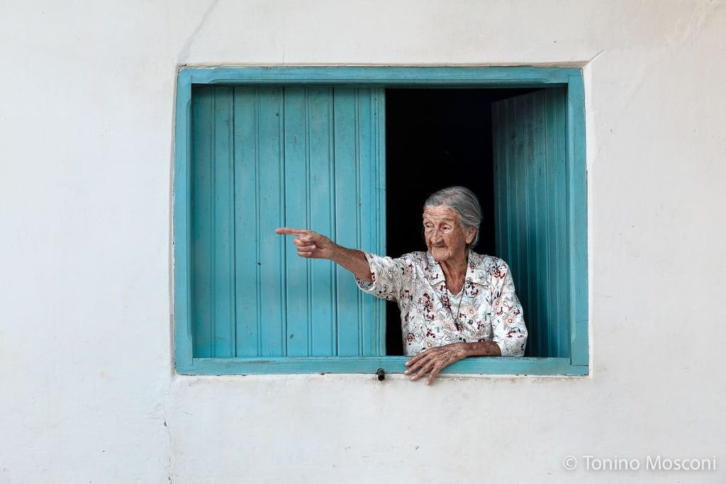 Fotografia di Tonino Mosconi di una donna anziana alla finestra, con persiane in legno azzurro, che indica qualcosa verso sinistra