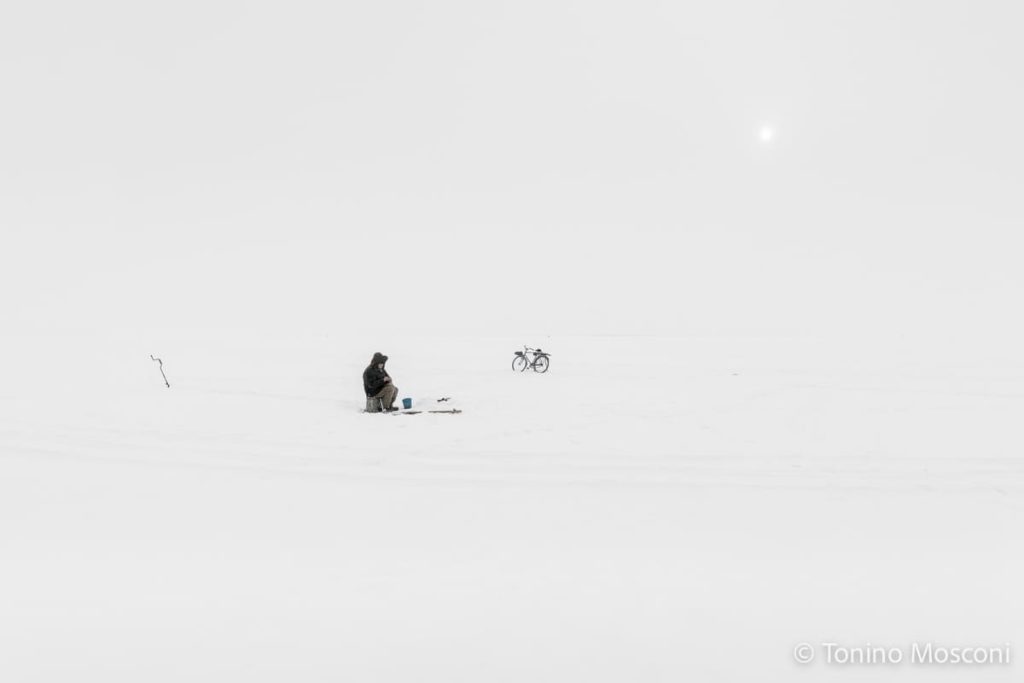 Fotografia di Tonino Mosconi ritraente un uomo nel mezzo di un distesa di neve, che si ferma e trova un momento di ristoro. Accanto a lui c'è la sua bicicletta.